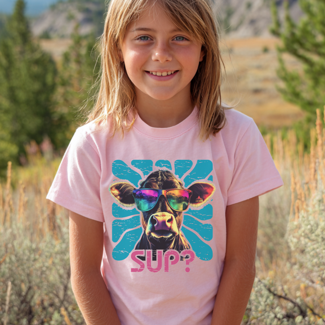 Young girl wearing a pink t-shirt with a cow graphic and 'SUP?' text in a natural setting.