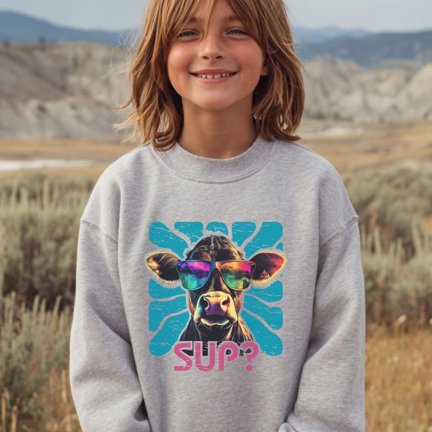 Child wearing a gray sweatshirt with a colorful cow graphic and 'SUP?' text in a desert landscape.