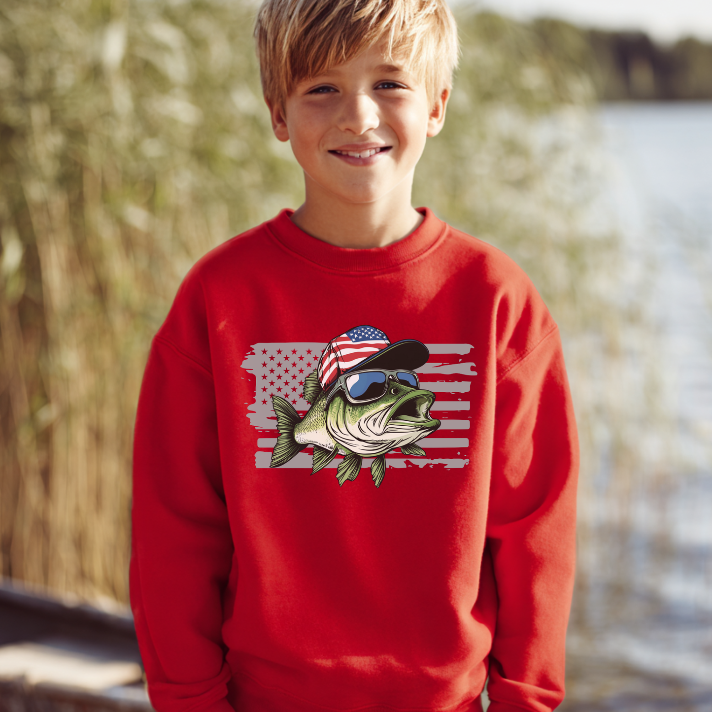 Child wearing a red sweatshirt with a fish design featuring an American flag and sunglasses, standing outdoors by water.