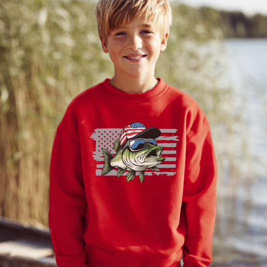 Child wearing a red sweatshirt with a fish design featuring an American flag and sunglasses, standing outdoors by water.