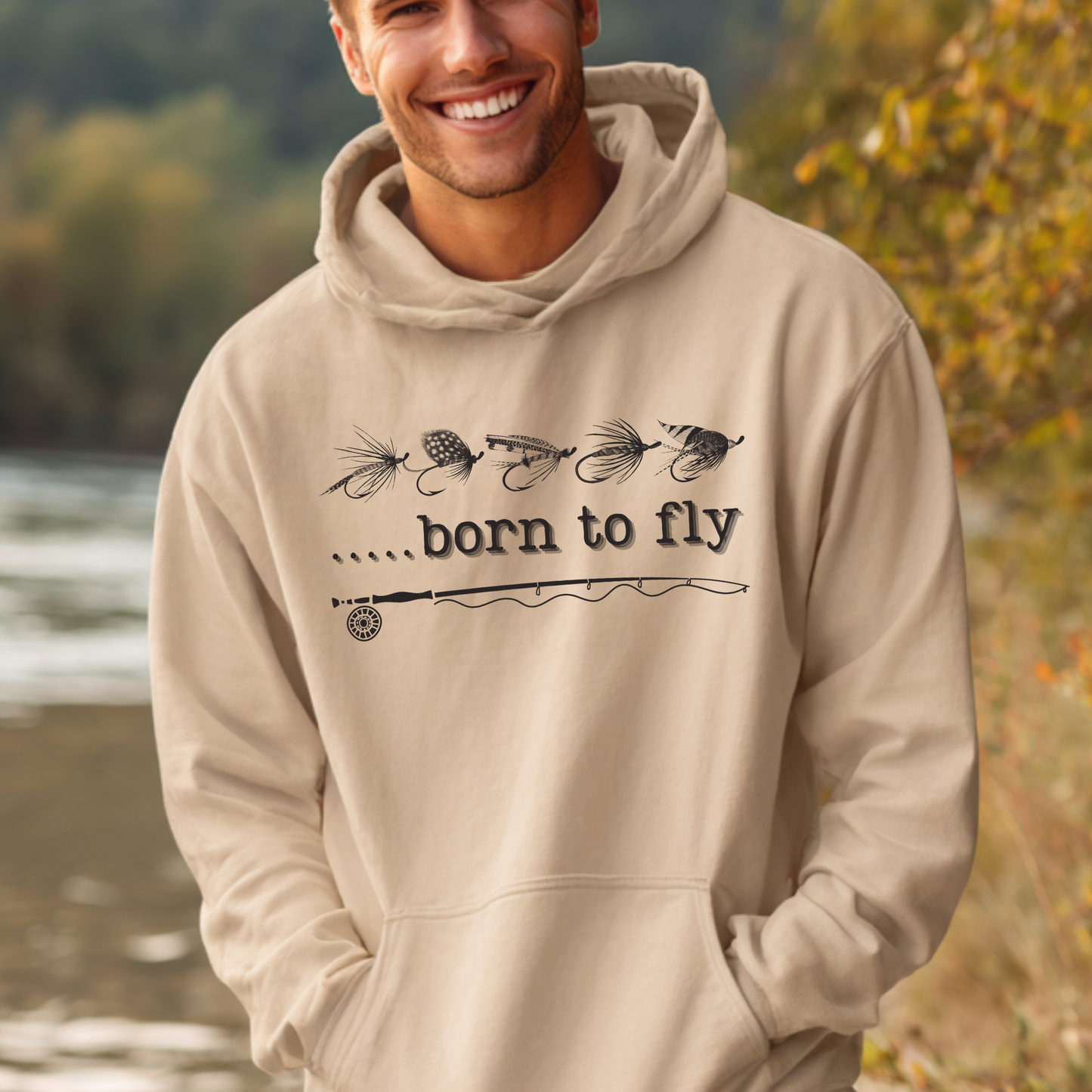 Man wearing a beige hoodie with 'born to fly' text and fishing lure designs outdoors.