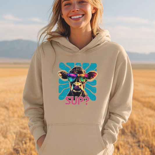 Person wearing a beige hoodie with a colorful cow graphic and 'SUP?' text, standing in a field with mountains in the background.
