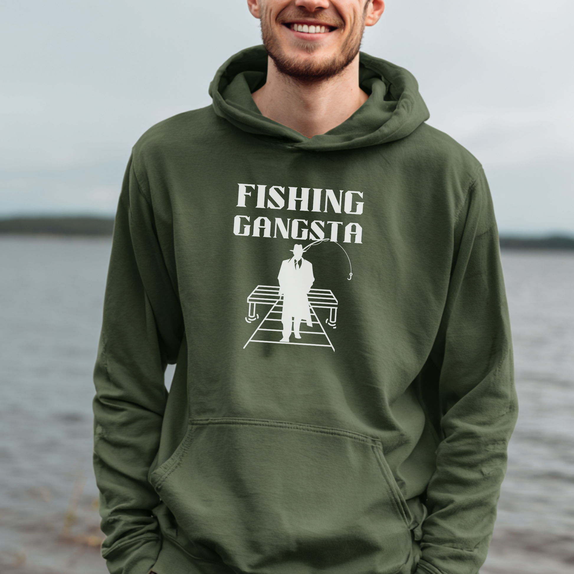Person wearing a green hoodie with 'Fishing Gangsta' graphic by a body of water.