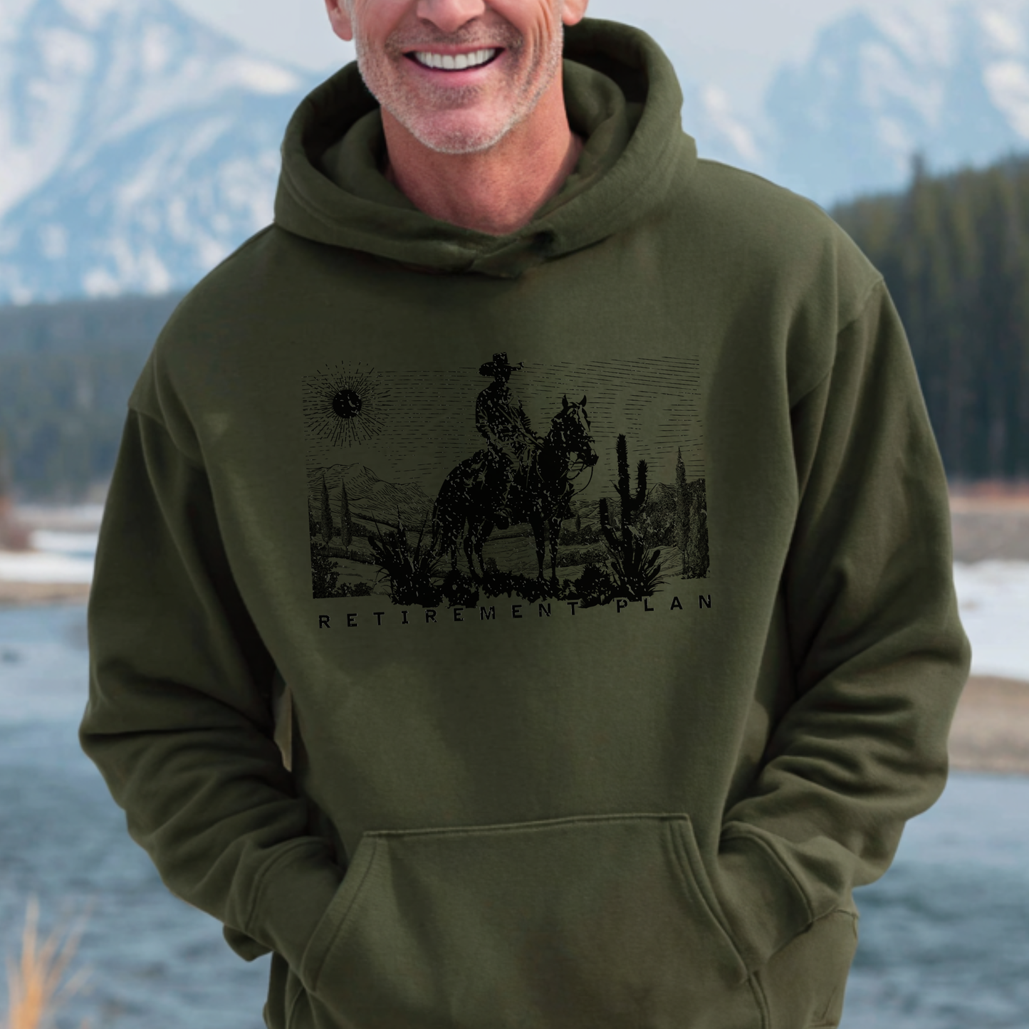 Man wearing a green hoodie with a graphic design of a person on a horse and text, standing in front of a scenic background.