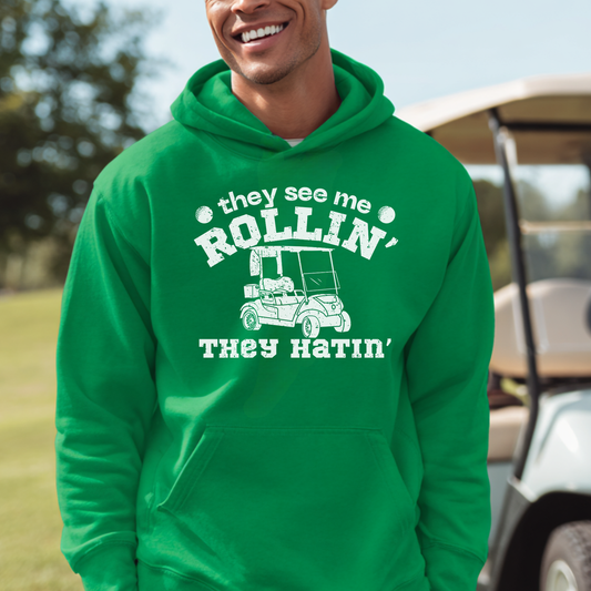 Person wearing a green hoodie with a humorous graphic and text, standing in a golf course setting.