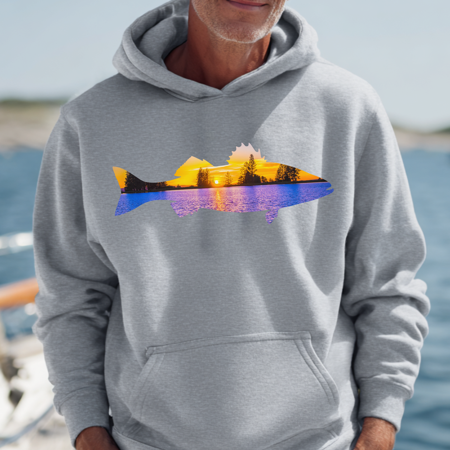 Person wearing a gray hoodie with a colorful mountain design by a body of water.