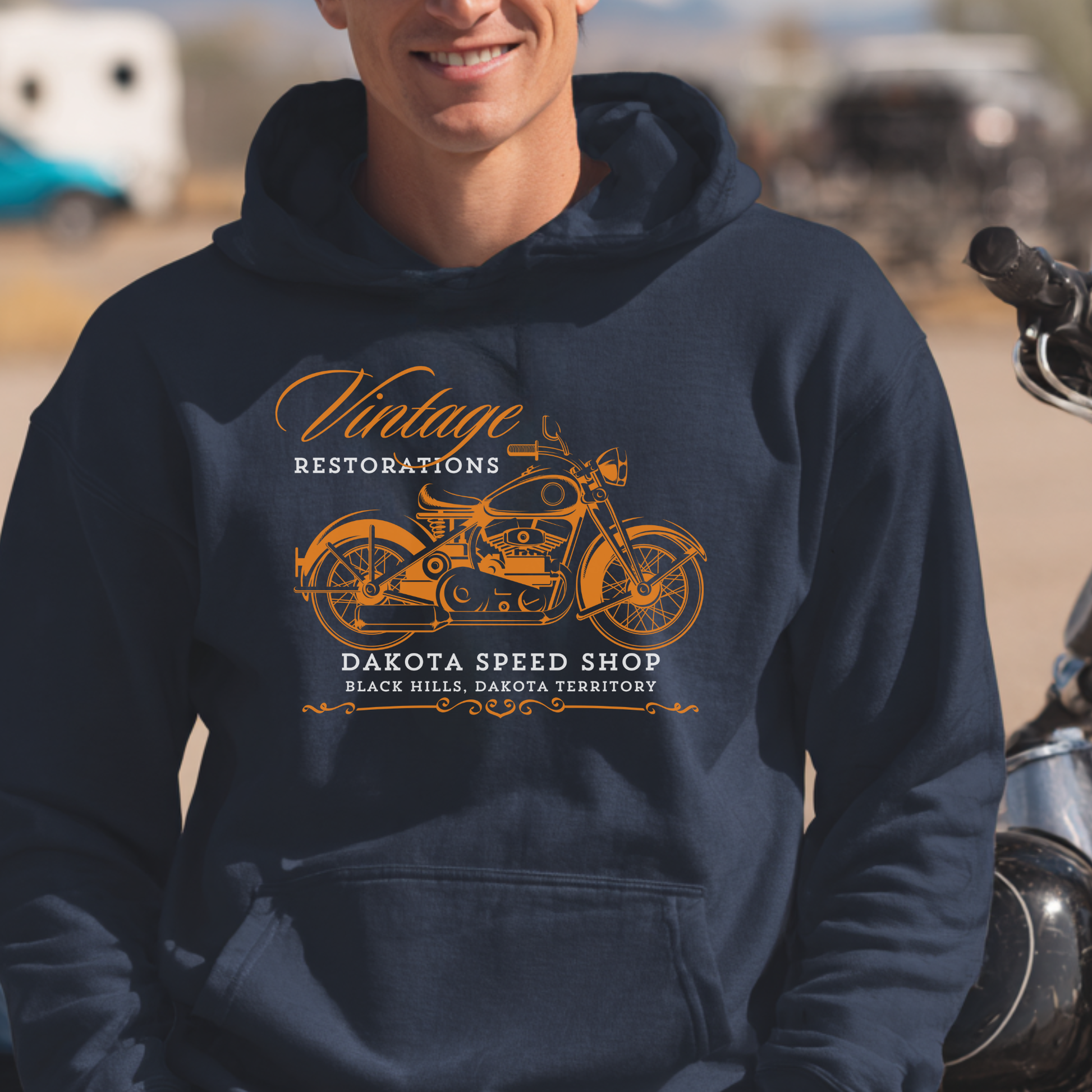 Person wearing a navy hoodie with a motorcycle graphic and text, standing next to a motorcycle.