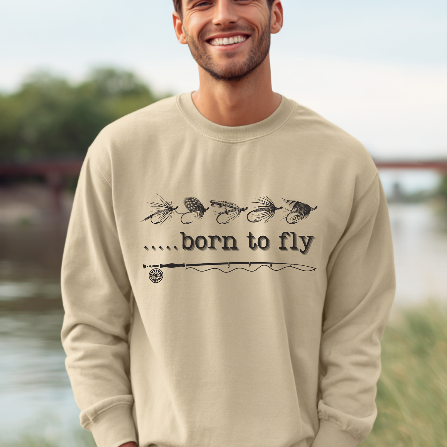 Man wearing a beige sweatshirt with 'born to fly' text and fishing lure graphics outdoors.