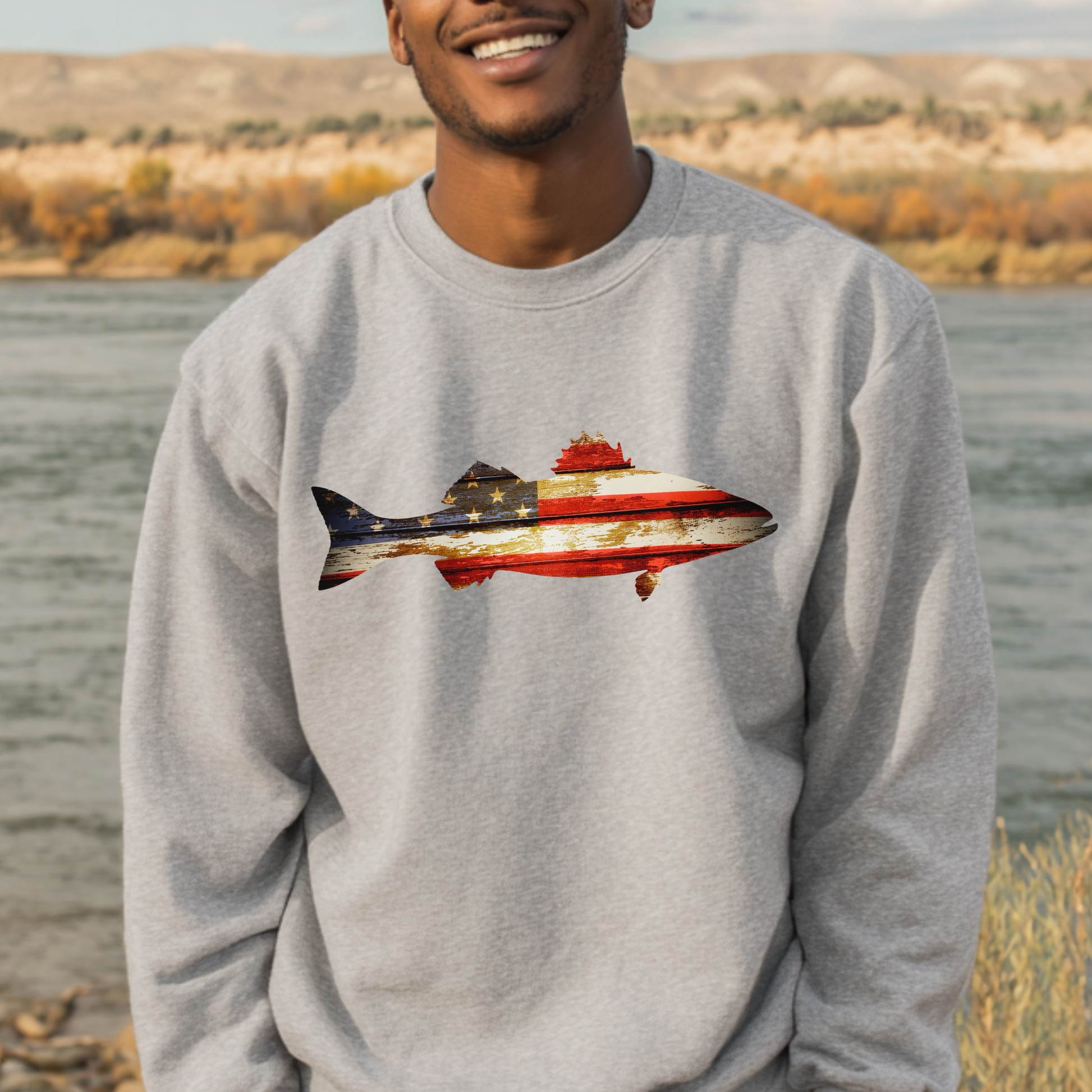 Person wearing a gray sweatshirt with a fish design featuring the American flag, standing by a body of water.