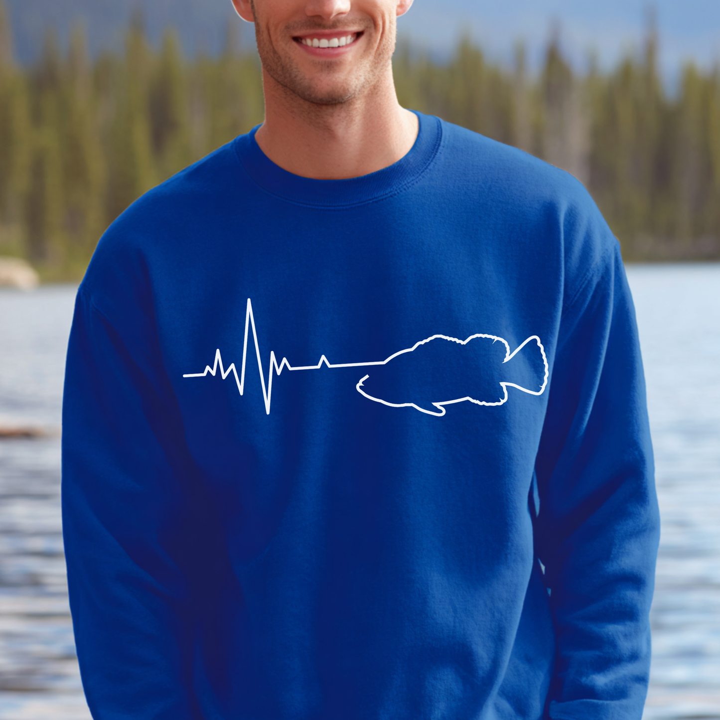 Man wearing a blue sweatshirt with a white fish and heartbeat line design, standing by a lake.
