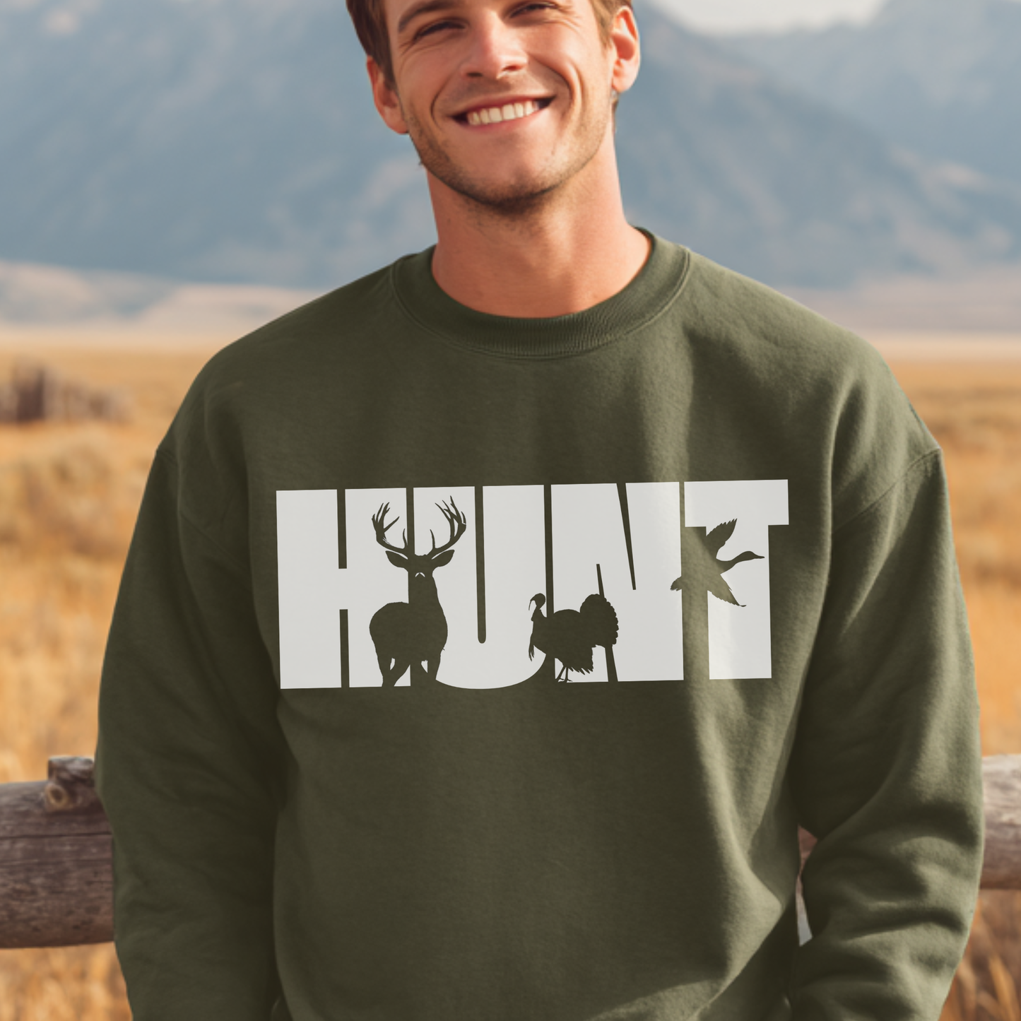 Man wearing a green sweatshirt with 'HUNT' and hunting-themed graphics against a mountainous background.