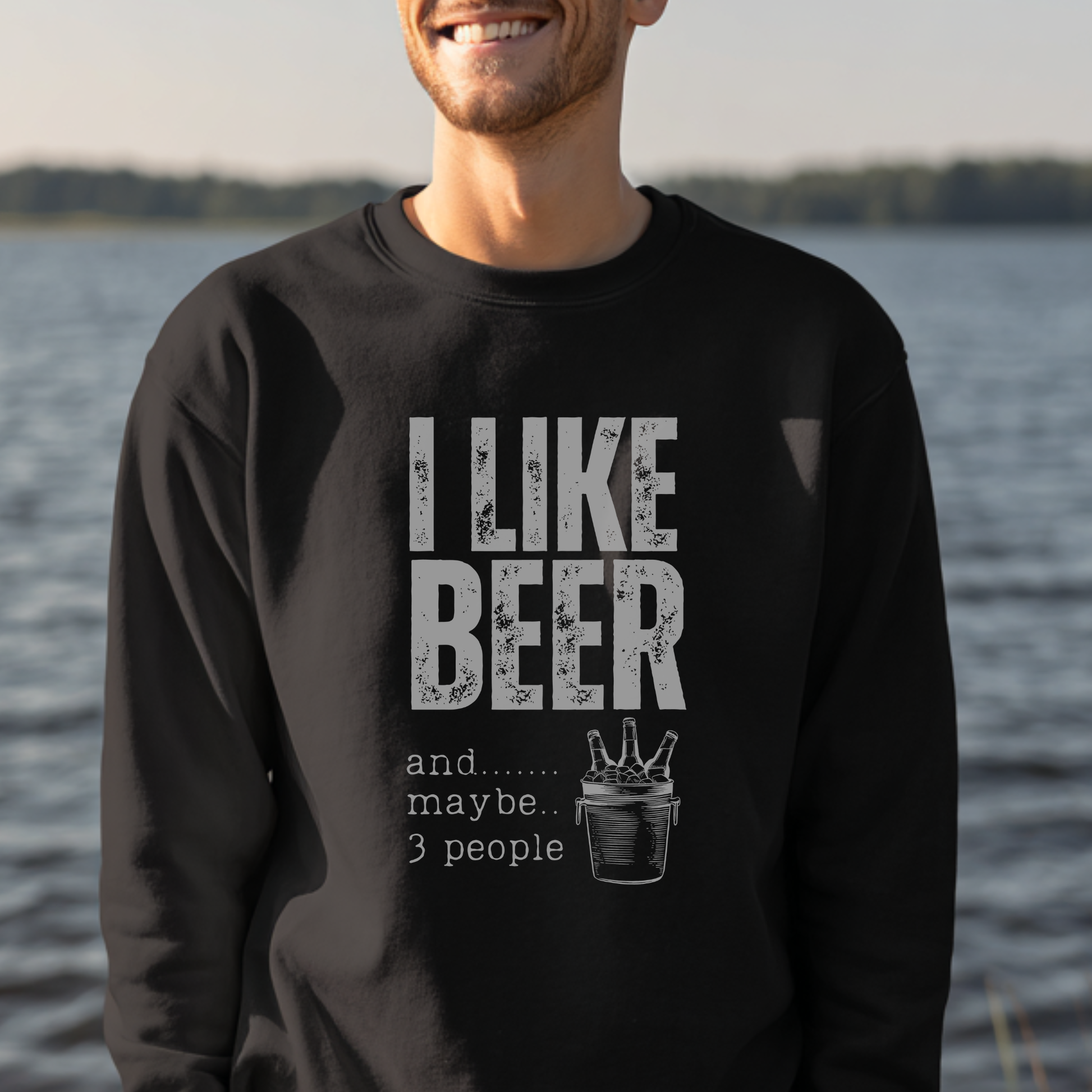 Person wearing a black sweatshirt with 'I LIKE BEER and...... maybe... 3 people' text by a body of water.