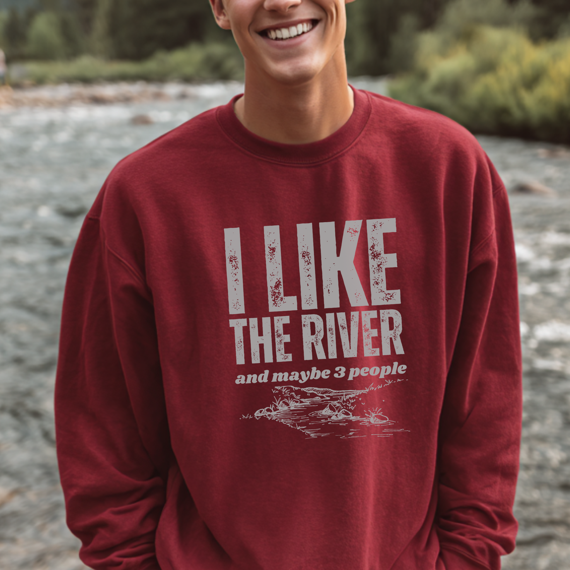Person wearing a red sweatshirt with 'I LIKE THE RIVER and maybe 3 people' text, standing by a river.