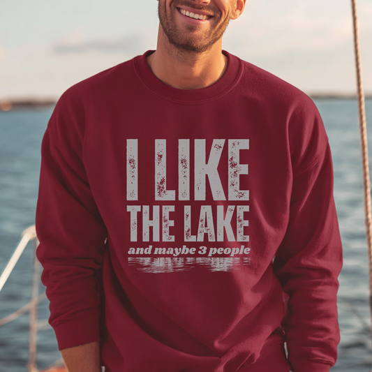 Man wearing a maroon sweatshirt with 'I Like the Lake and maybe 3 people' text by a lake.