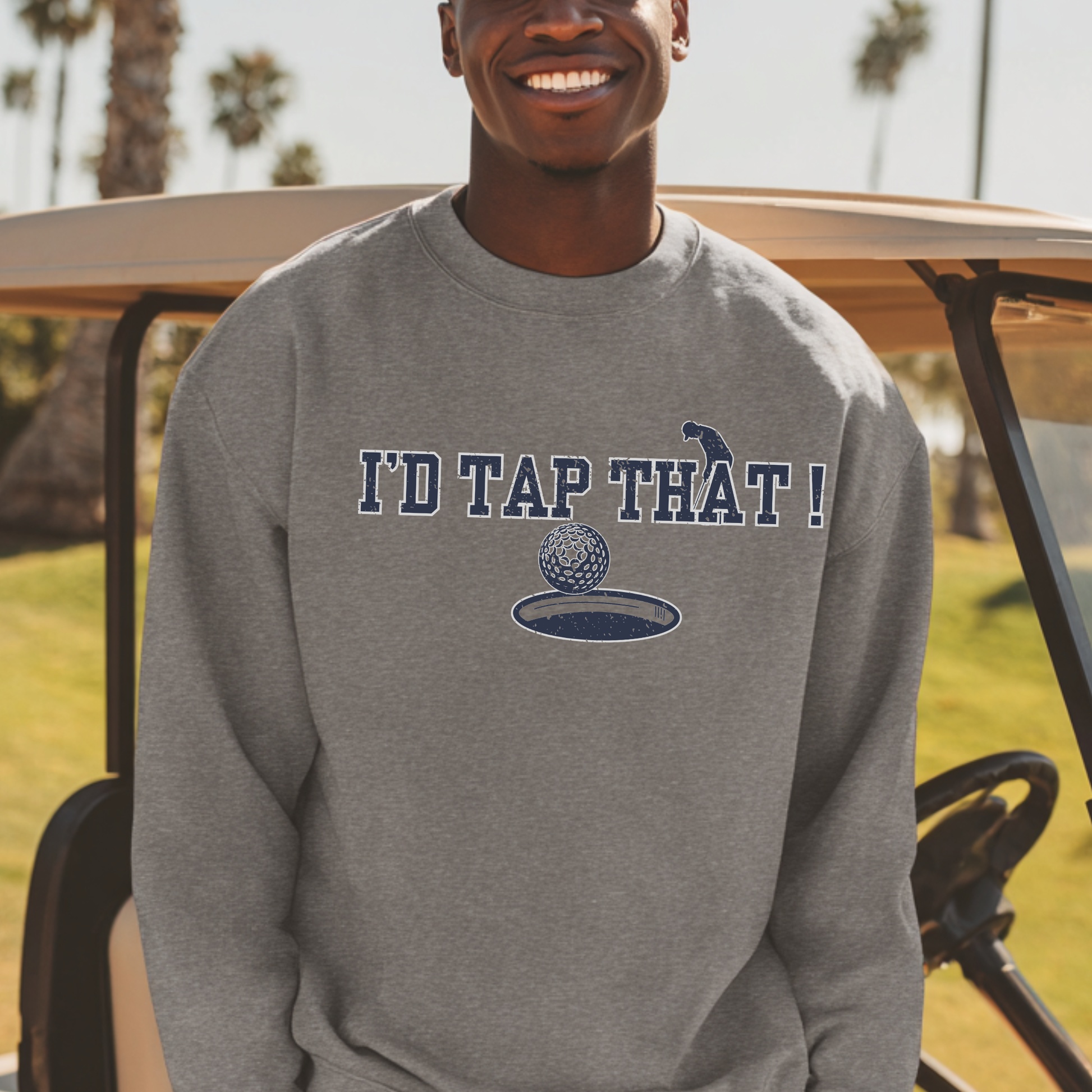 Person wearing a gray sweatshirt with 'I'd Tap That!' text and graphic, standing in front of a golf cart on a golf course.