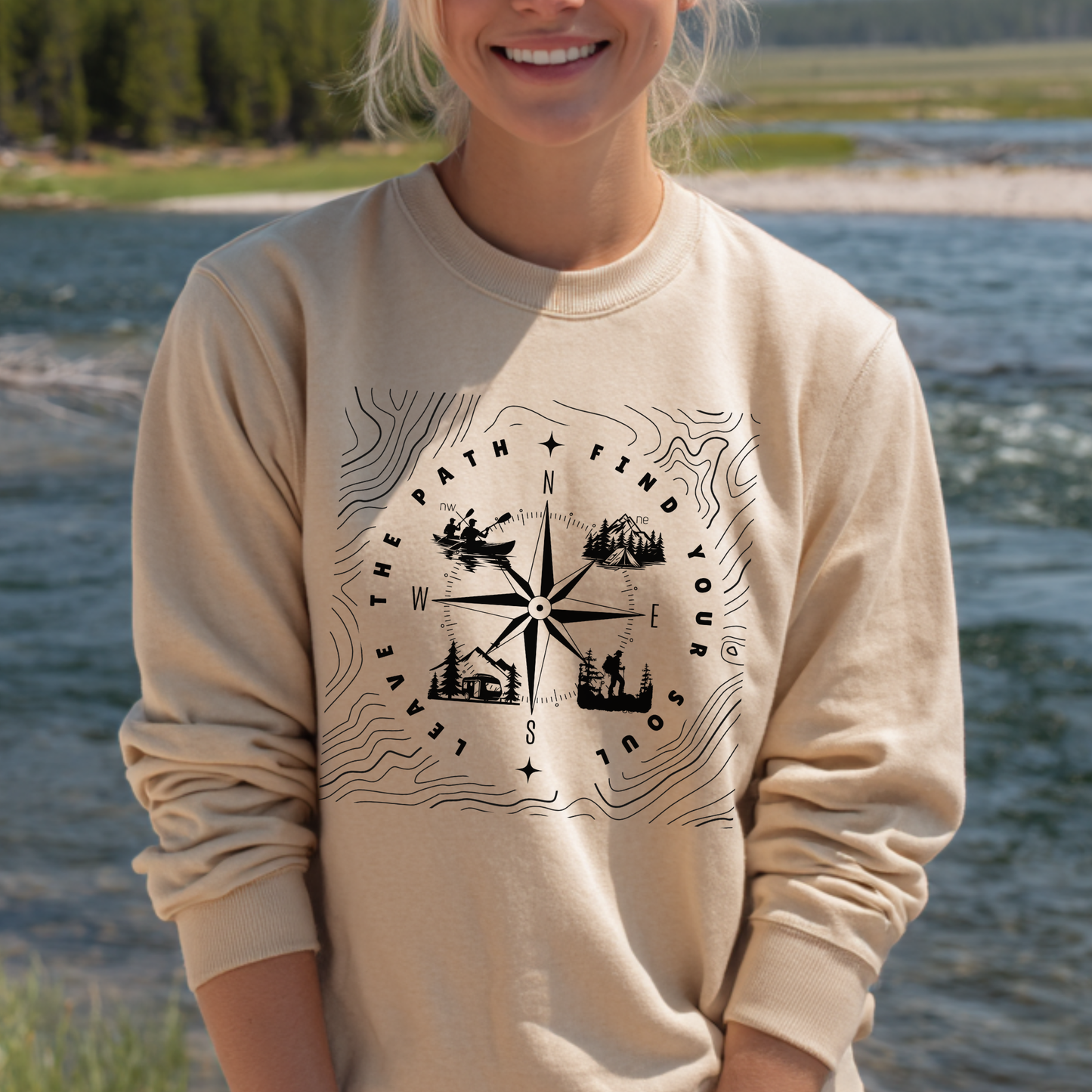 Person wearing a beige sweatshirt with a compass design by a body of water