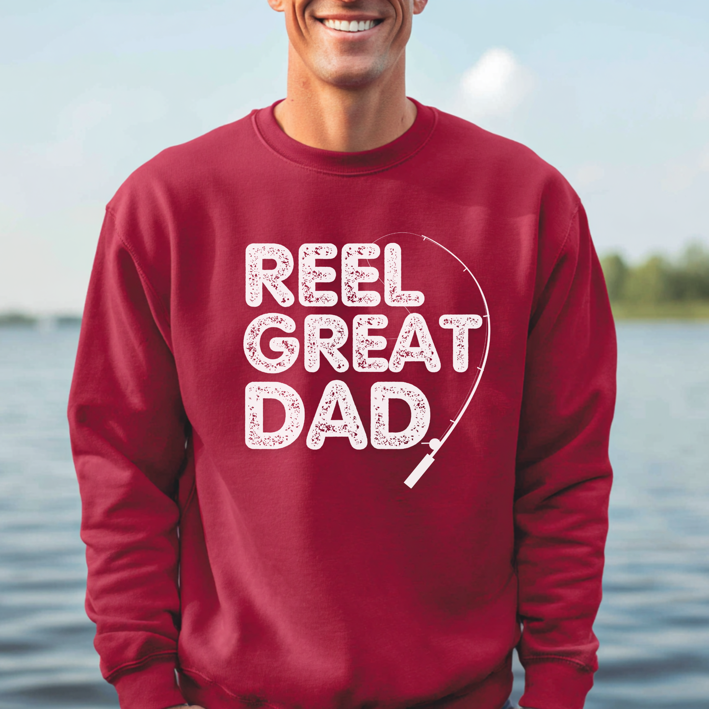 Person wearing a red sweatshirt with 'REEL GREAT DAD' text by a body of water.
