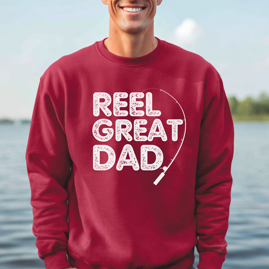 Person wearing a red sweatshirt with 'REEL GREAT DAD' text by a body of water.