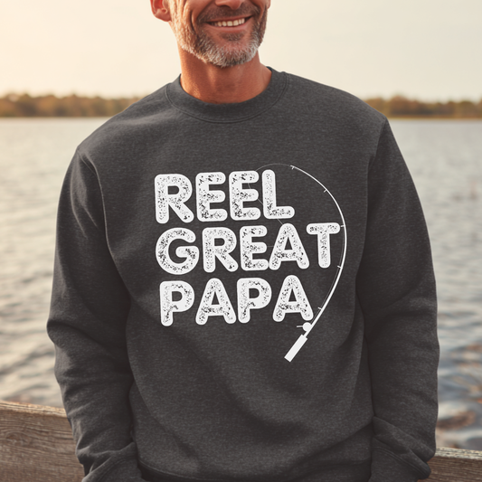 Man wearing a dark gray sweatshirt with 'Reel Great Papa' text by a body of water.