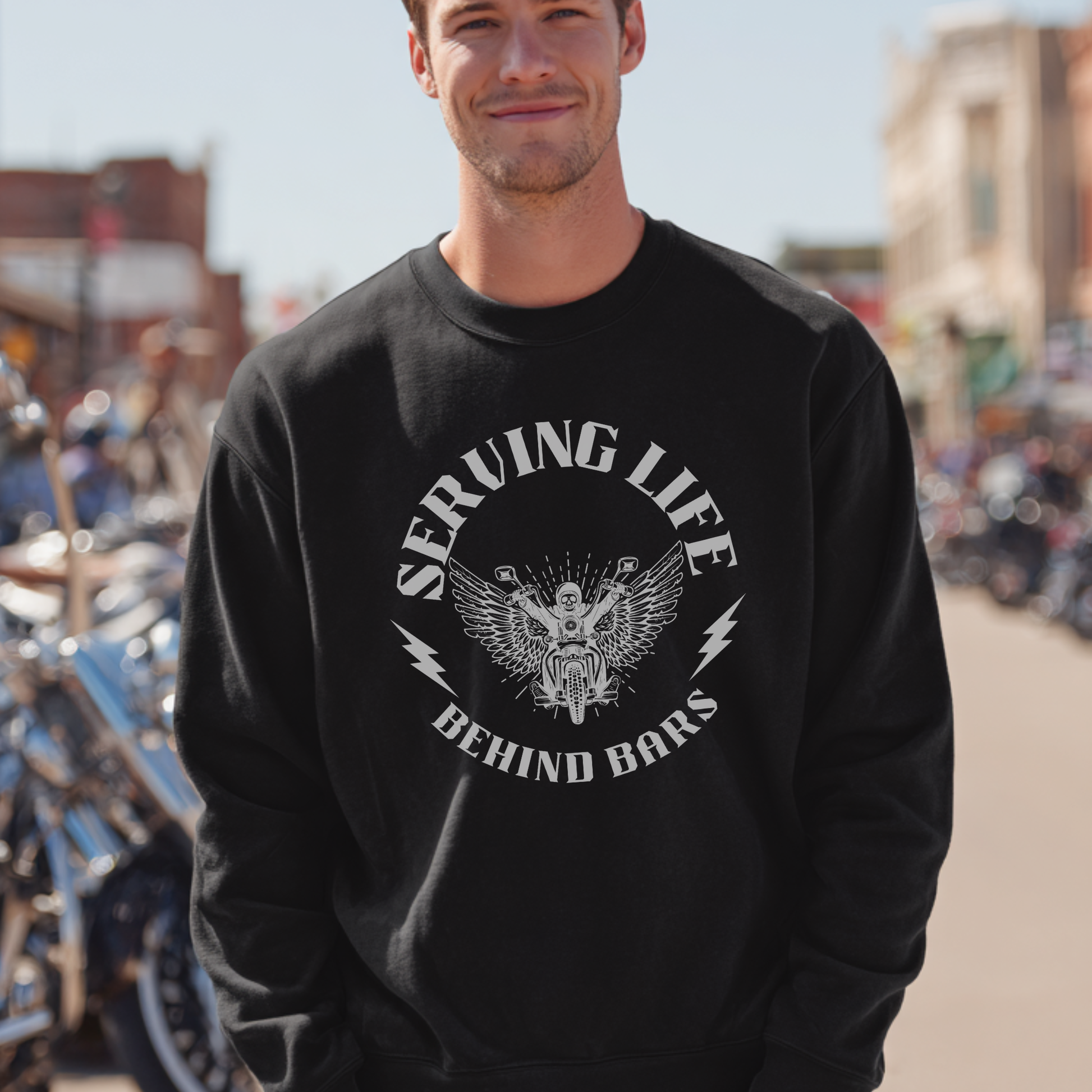 Man wearing a black sweatshirt with a graphic design and text, standing in an outdoor setting with motorcycles.