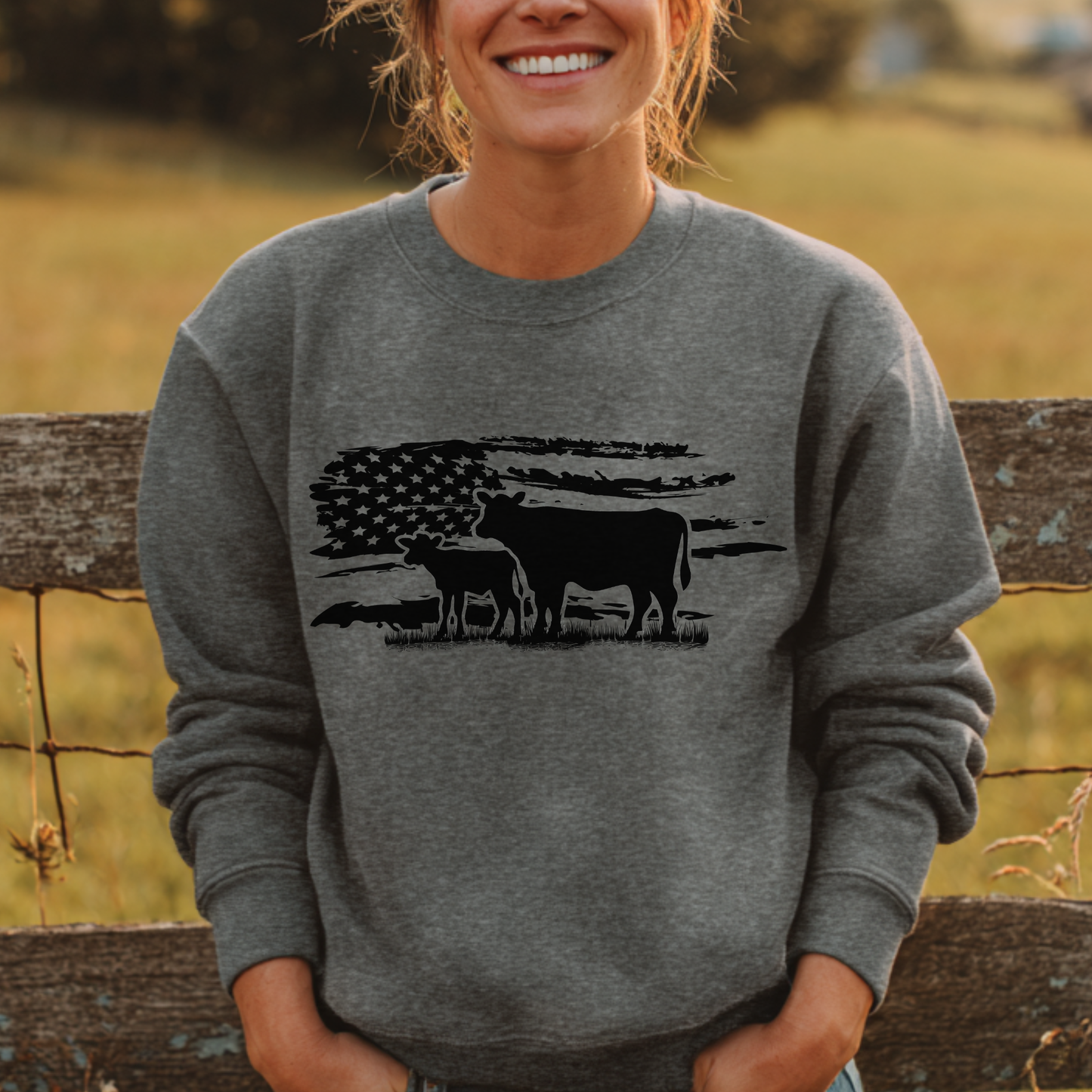 Person wearing a gray sweatshirt with a cow and flag design, standing in front of a wooden fence.