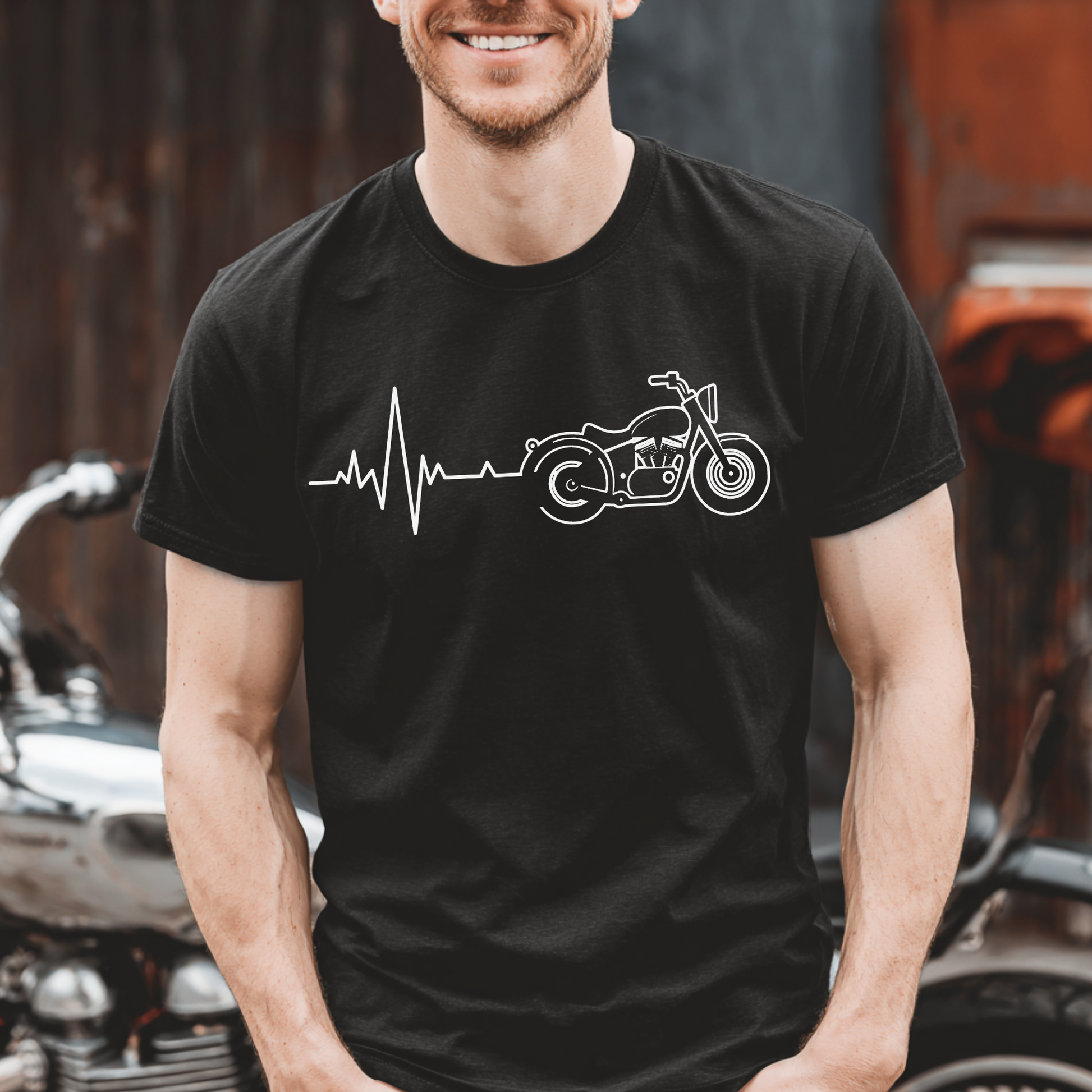 Man wearing a black t-shirt with a motorcycle and heartbeat line design, standing in front of a motorcycle.