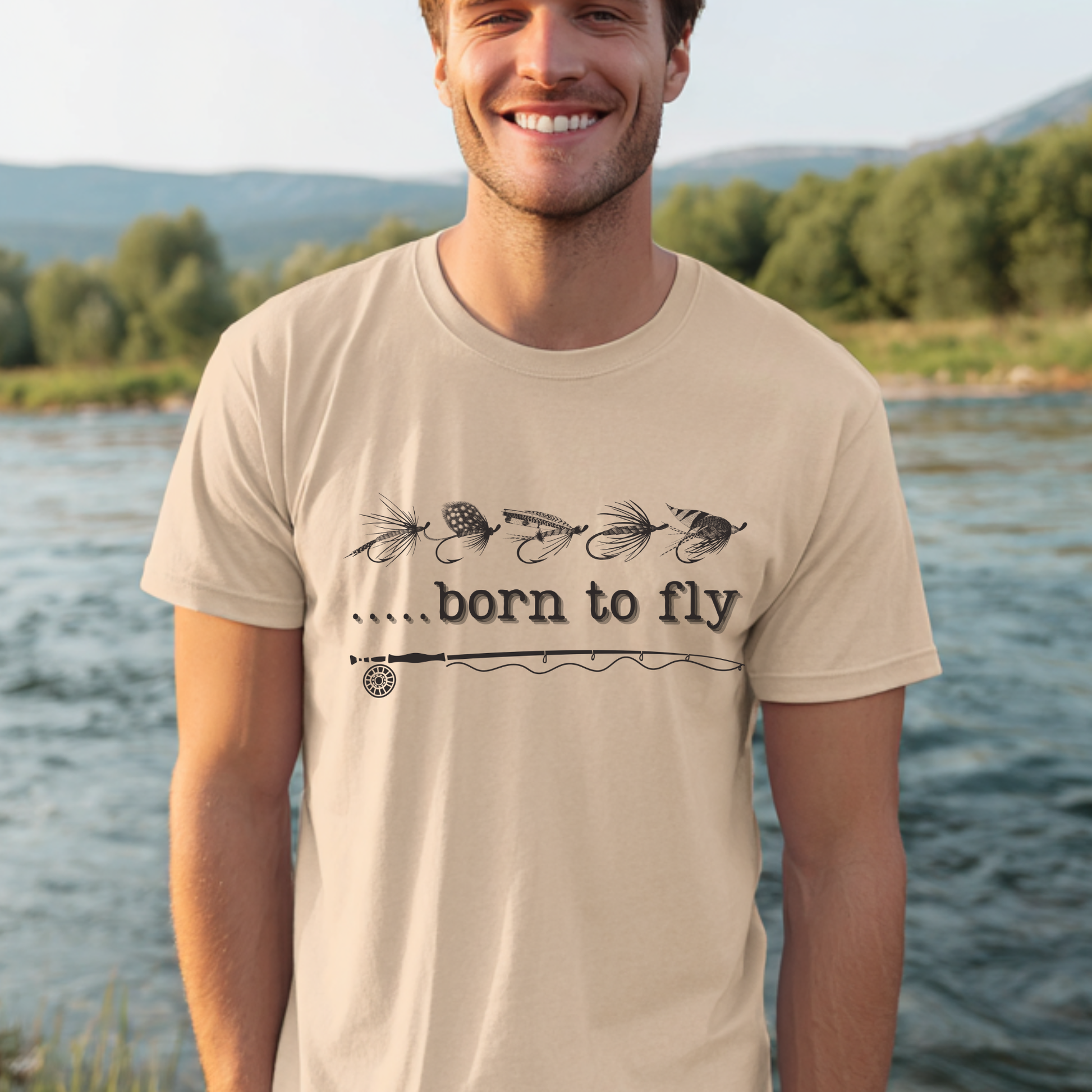 Man wearing a beige t-shirt with 'born to fly' text and fishing lure graphics by a lake.