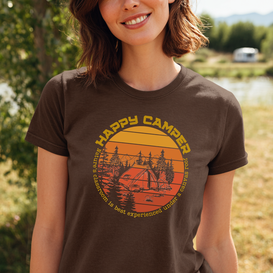 Person wearing a 'Happy Camper' t-shirt with a sunset and camping design outdoors.