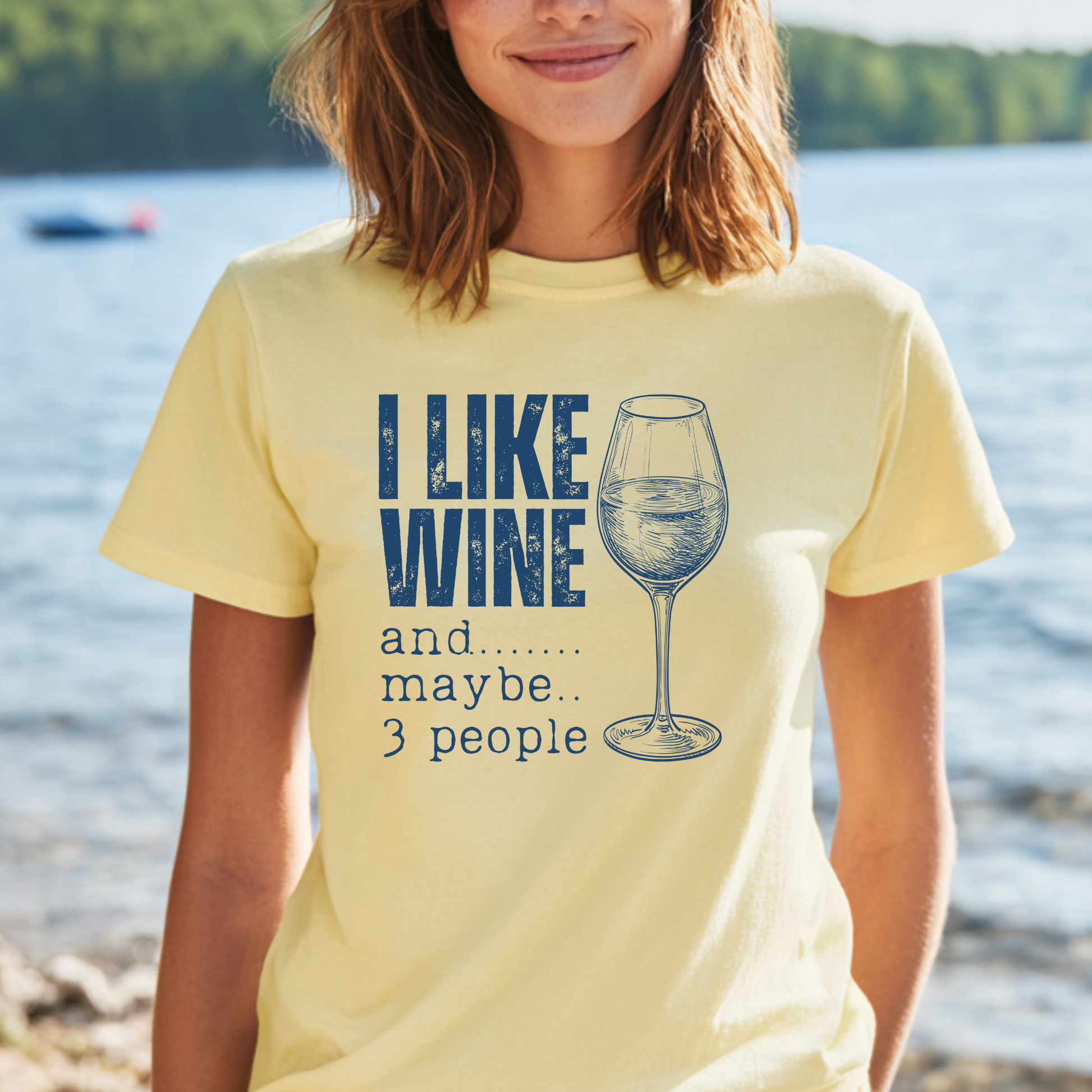 Person wearing a yellow t-shirt with a wine-themed design by a lake.