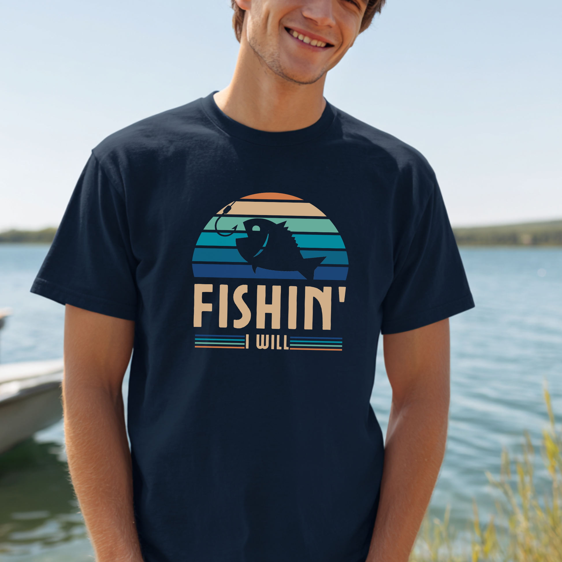 Person wearing a black t-shirt with 'FISHIN' I WILL' graphic by a body of water.
