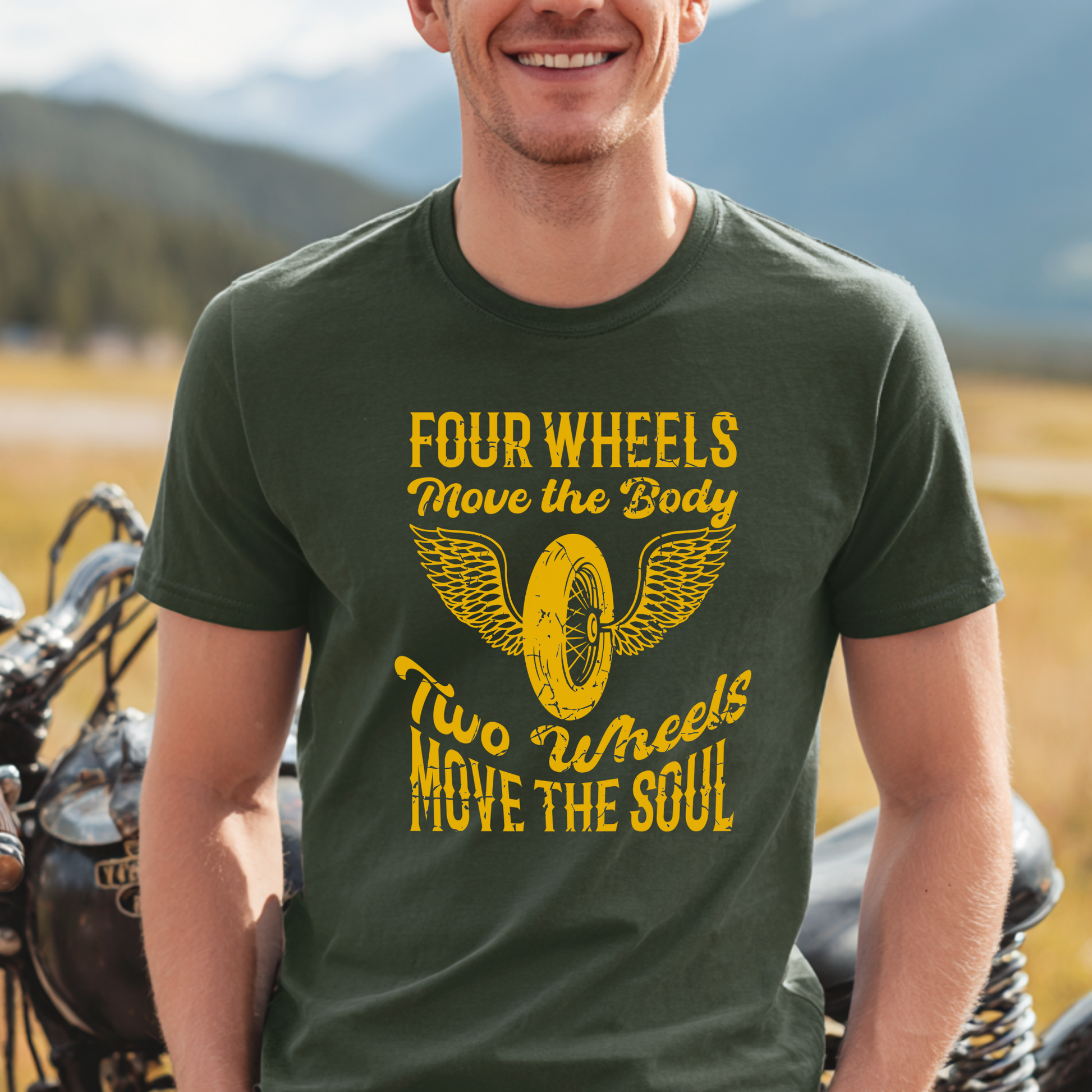 Man wearing a green t-shirt with a yellow motorcycle graphic and text, standing next to a motorcycle with a mountainous background.