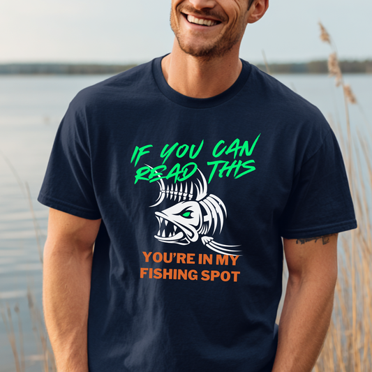 Man wearing a navy blue t-shirt with a humorous fishing graphic and text by a body of water.