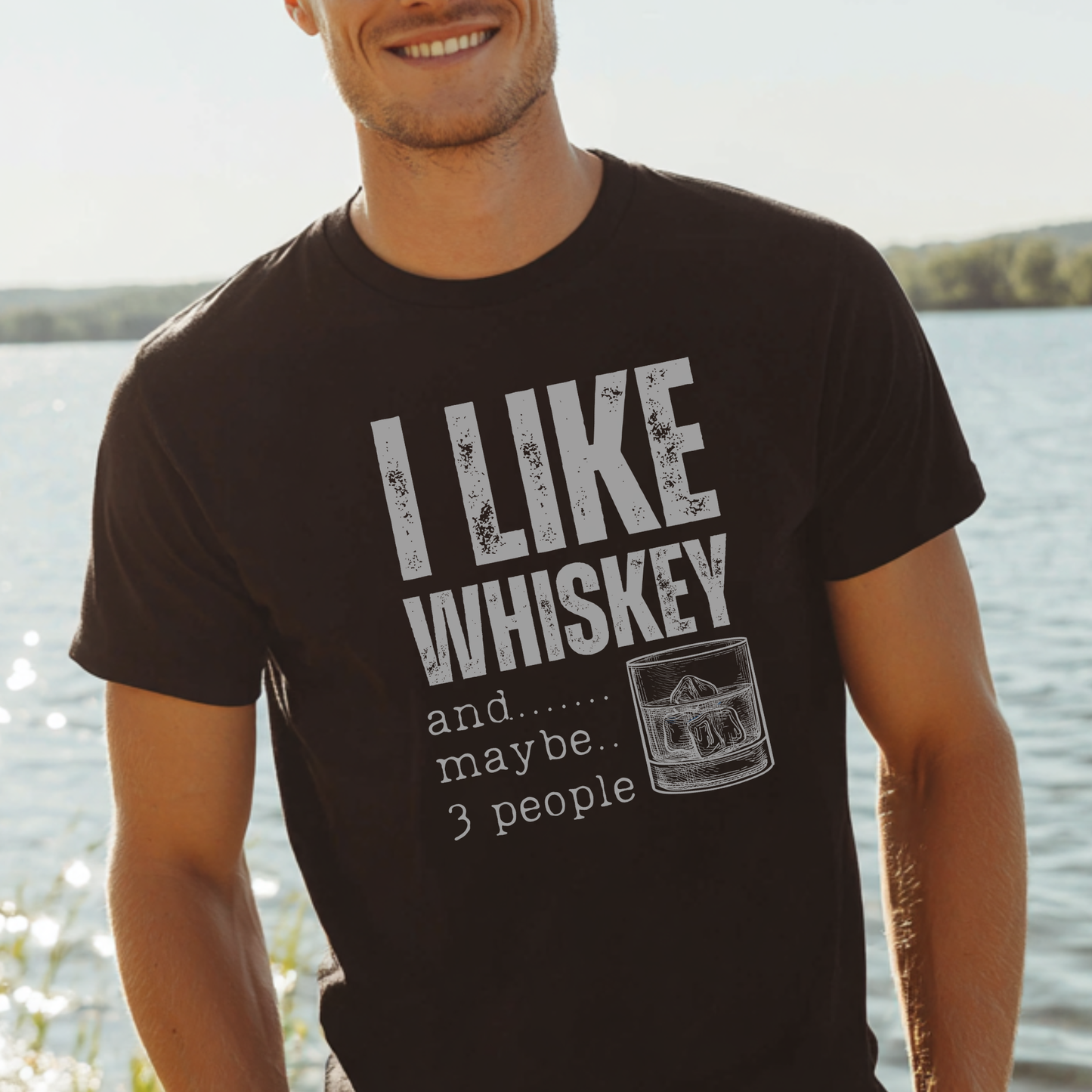 Man wearing a black t-shirt with 'I Like Whiskey and maybe 3 people' text by a body of water.