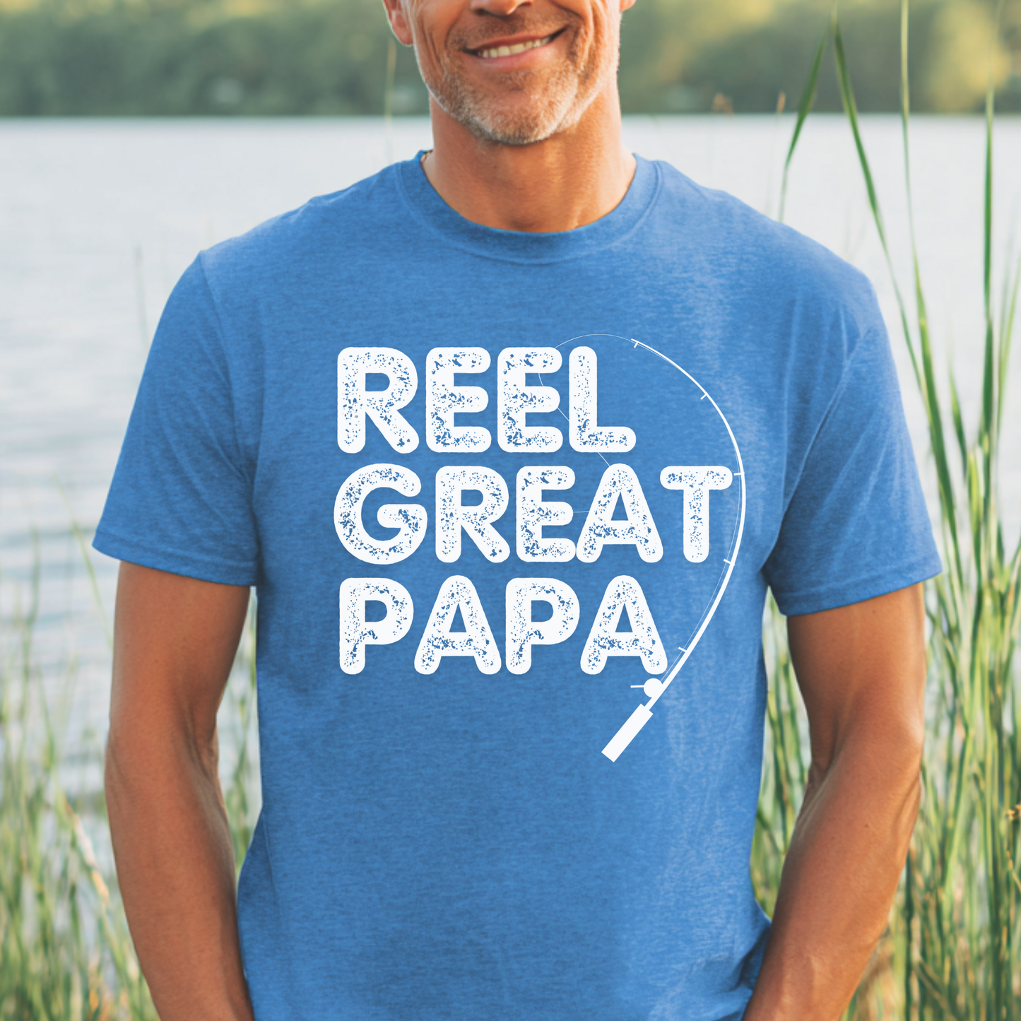 Man wearing a blue t-shirt with 'Reel Great Papa' text by a body of water.