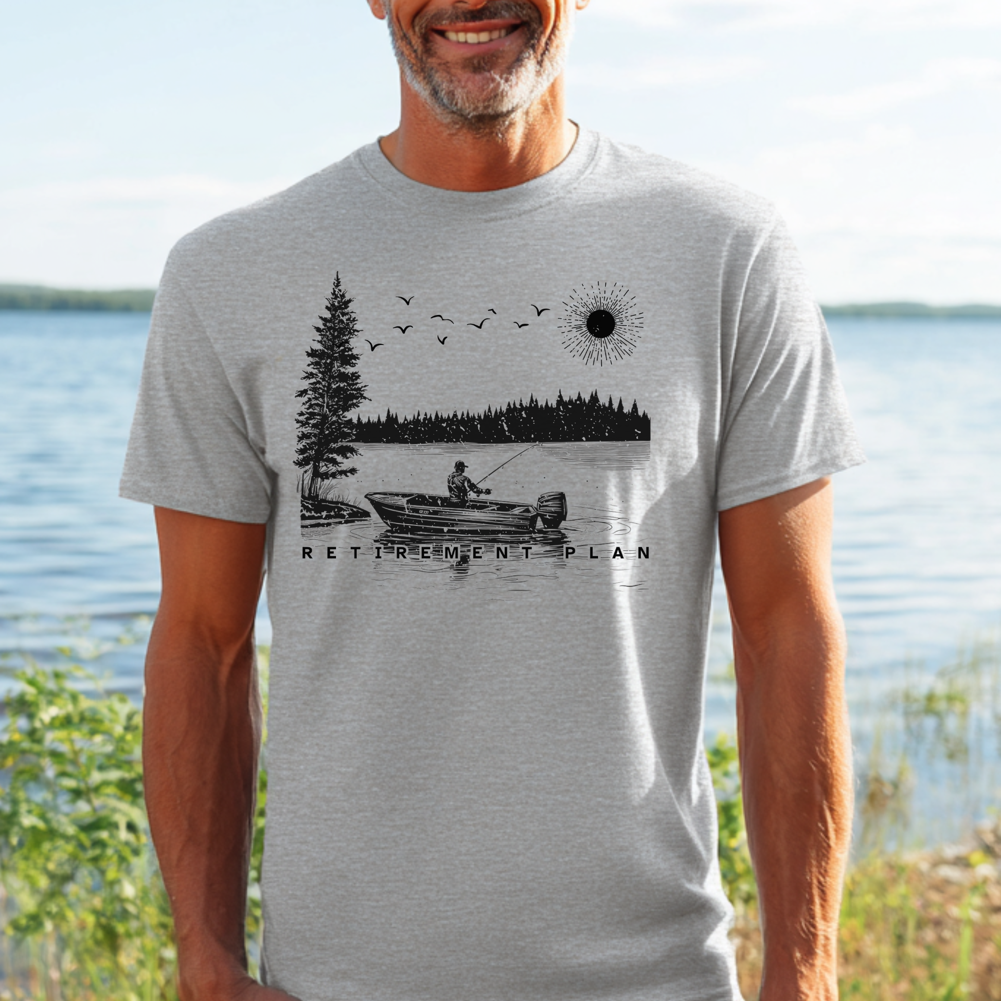 Man wearing a t-shirt with a fishing graphic and 'Retirement Plan' text by a lake.