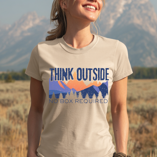 Person wearing a 'Think Outside No Box Required' t-shirt with a mountain design in a natural setting.