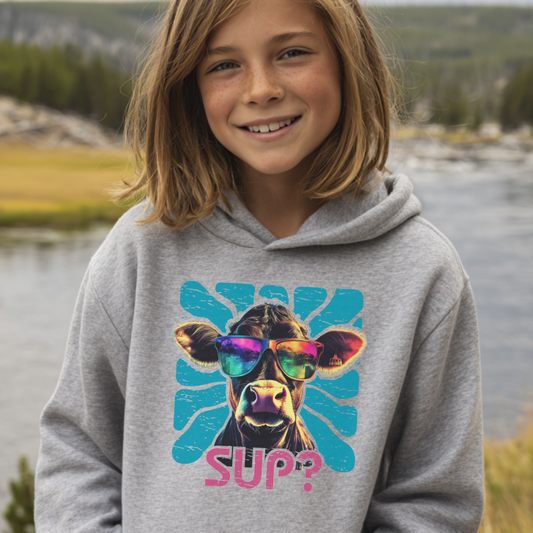 Child wearing a hoodie with a cow graphic and 'SUP?' text, standing outdoors by a body of water.