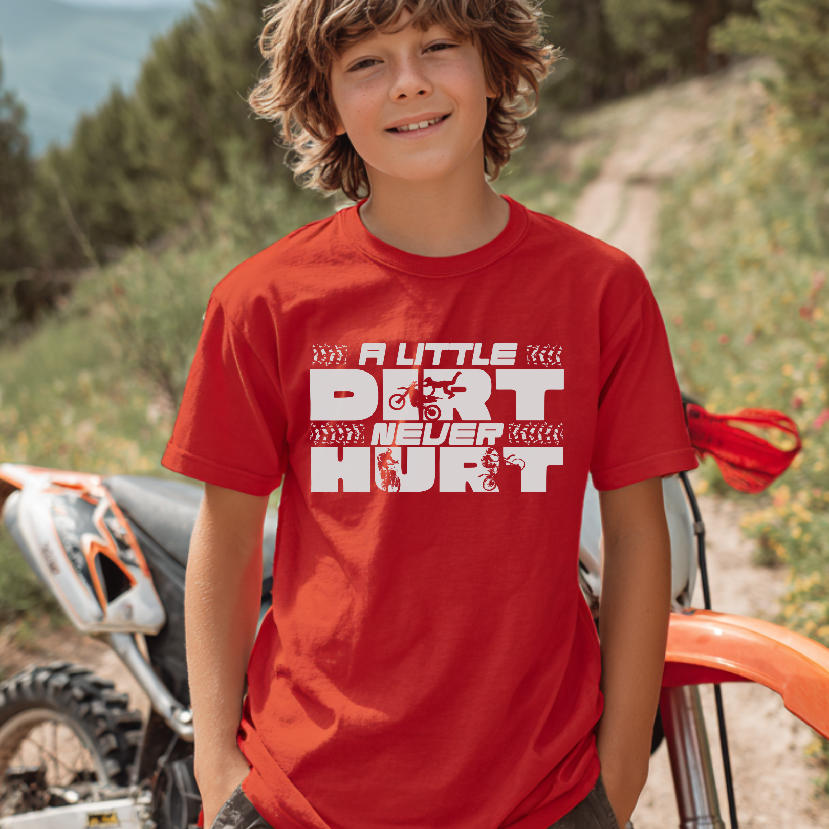 Child wearing a red t-shirt with text standing next to a dirt bike on a trail.