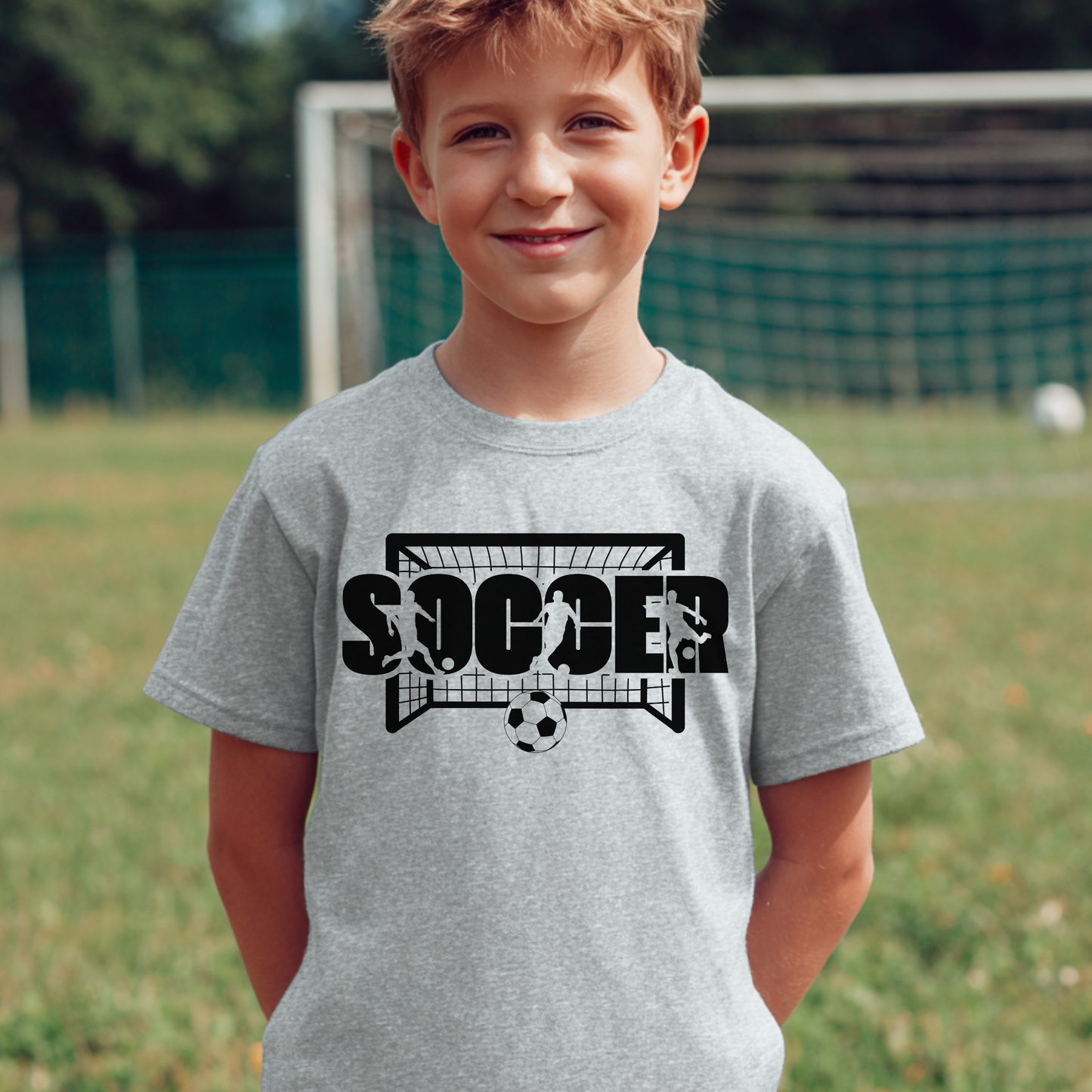 Child wearing a gray t-shirt with a soccer graphic and text on a grassy field.